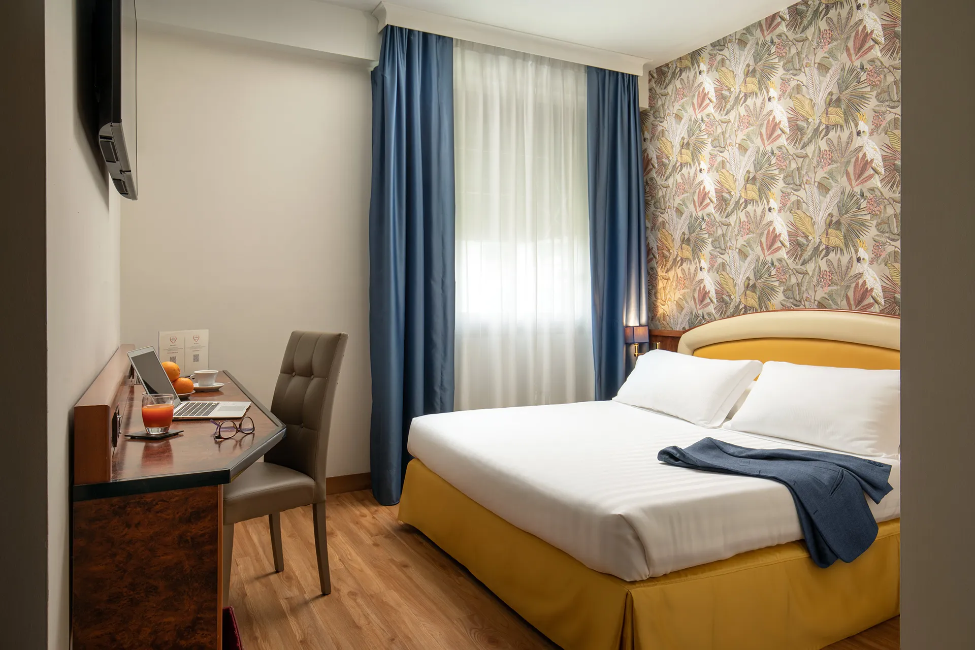 In the picture, the Business Room at the Park Hotel Roma Nord: we can see the double bed against a wall with floral wallpaper. A jacket is on the bed. O the other sideof the room, a desk with a laptop, a cup of coffee, a glass of juice and a pair of glasses.