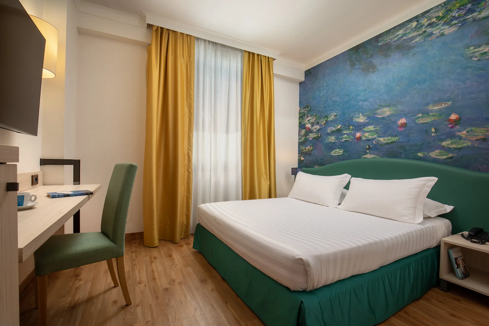 In the picture, the Executive Room of the Park Hotel Roma Nord: we can see the double bed and, on the other side of the room, a table and a TV hanging on the wall. Behind the bed, a wallpaper with water lilies in a pond.