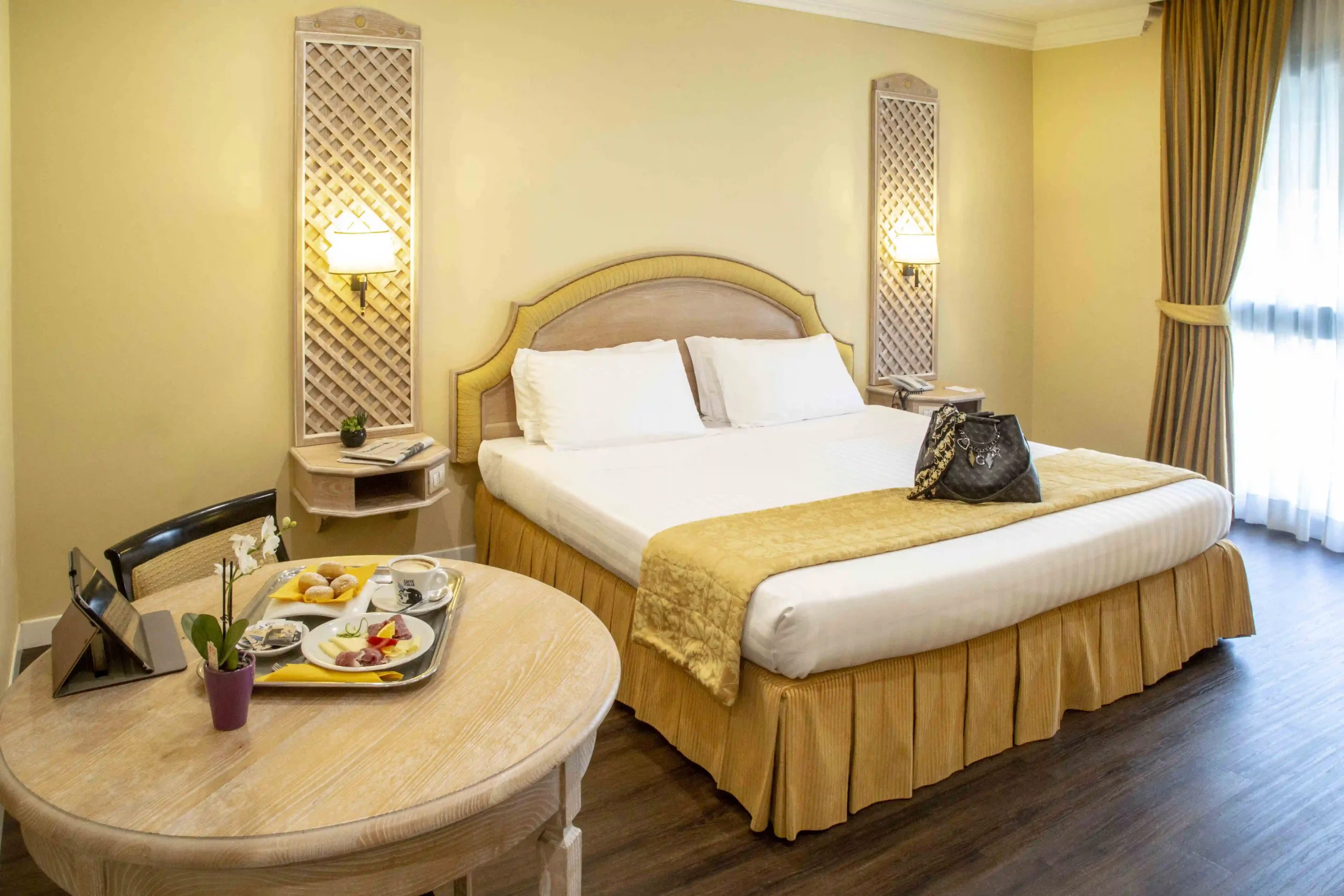 In the picture, the Suite at the Park Hotel Roma Nord: we can see the double bed, on which a bag is placed. On the left, a small round table with a tablet, a tray with a plate of cured meats and cheese and a cappuccino.