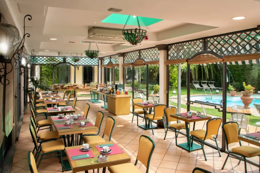 In the picture, the Breakfast Room at the Park Hotel Roma Nord: inside a veranda overlooking the pool, there are some tables and, on the left, the breakfast buffet.