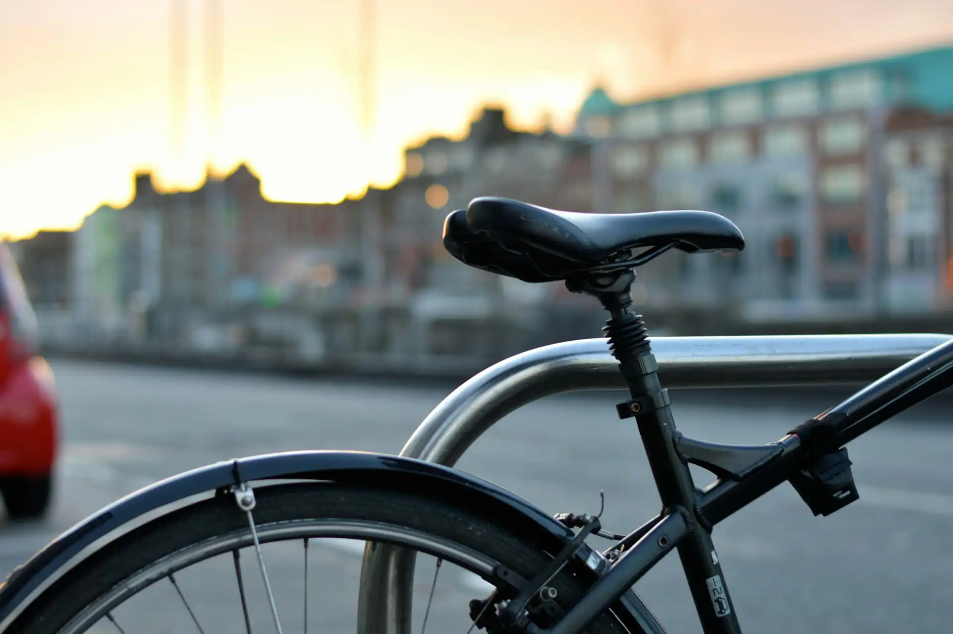 In the picture, a close-up of the saddle of a black bicycle. In the background, we can see some buildings out of focus.
