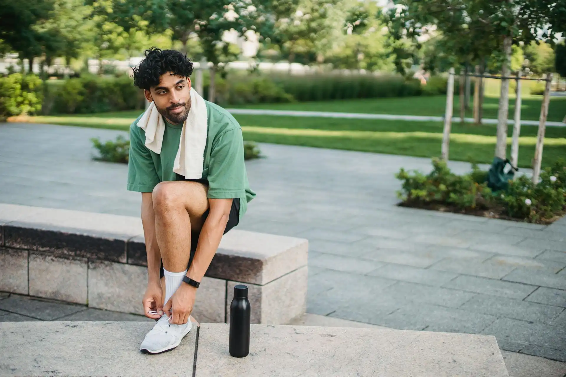 In the picture, a man in athletic gear tying his shoelaces with a towel draped over his shoulders. A black water bottle rests beside him. In the background, a park.